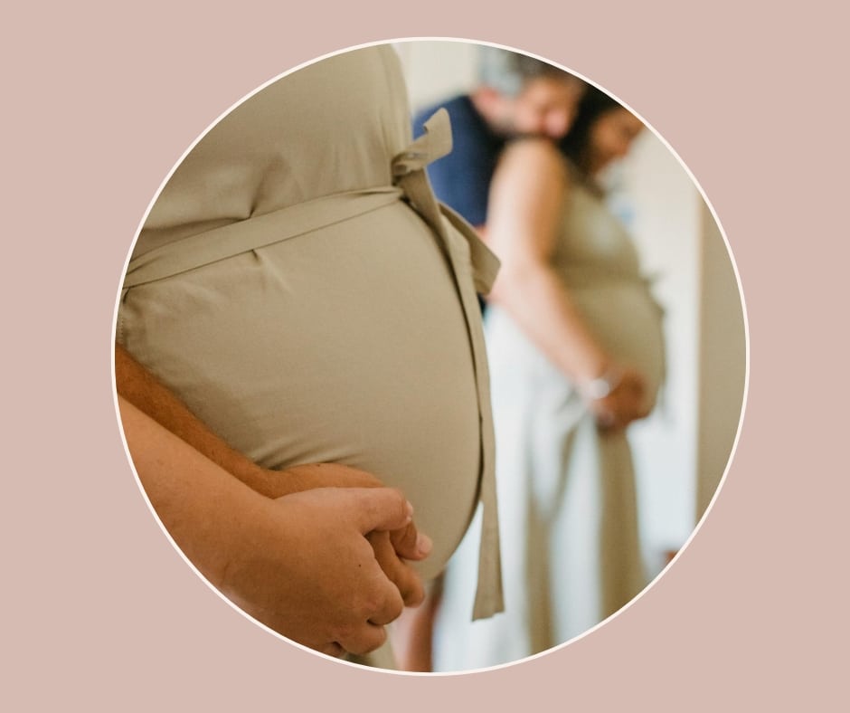 Nahaufnahme eines schwangeren Bauchs mit sanft gefalteten Händen, im Hintergrund weitere Schwangere – Szene aus einem Geburtsvorbereitungskurs.